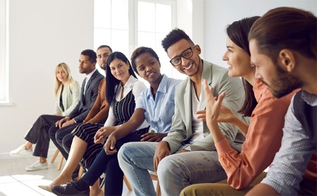 A diverse group of young adults seated in a line of chairs chats happily