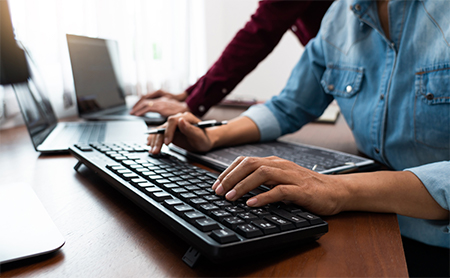 midsection shot of a desk with a man in the foreground using a full-size keyboard, the woman behind him is using a laptop