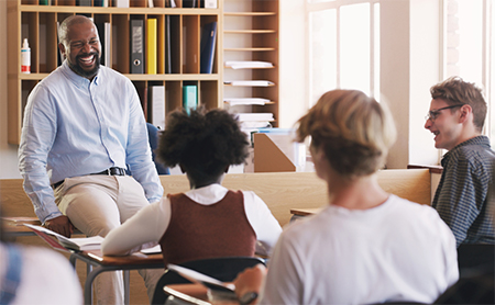 A teacher sits on his desk, facing students, as they all laugh together