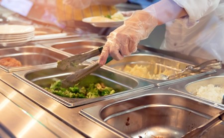 A worker serving food from behind full lunch service station in a cafeteria.